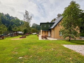 A building with a green roof and beige walls, surrounded by a grassy area with picnic tables and a wooden deck. - Apartment Meda by Interhome (Rudnik)