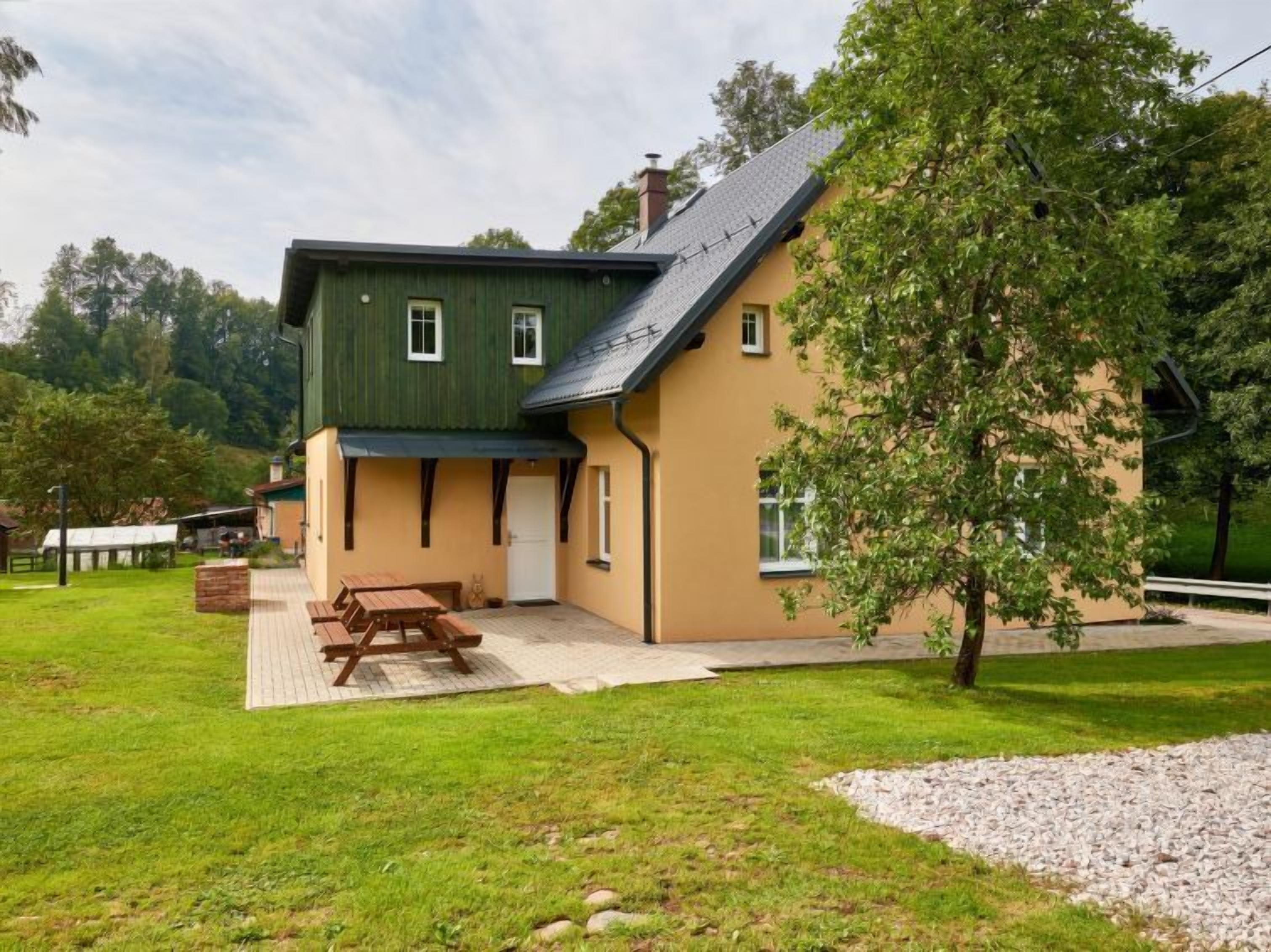Kaksikerroksinen talo, jossa on vihreä katto, beigen sävyiset seinät ja puinen terassi, jossa on piknikpöytiä.