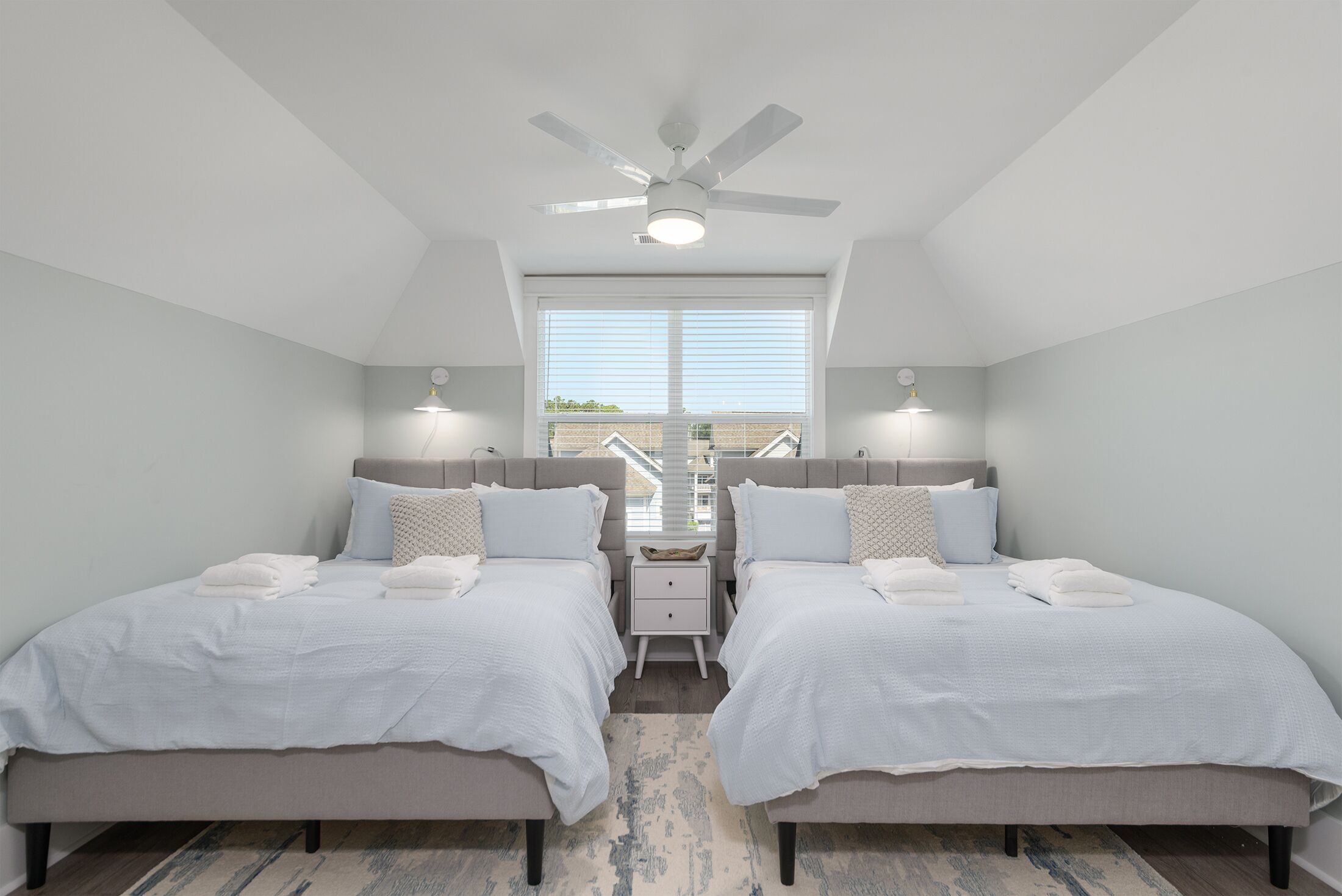 A twin bedroom with two beds, a ceiling fan, bedside lamps, a window with blinds, and a view of buildings outside.
