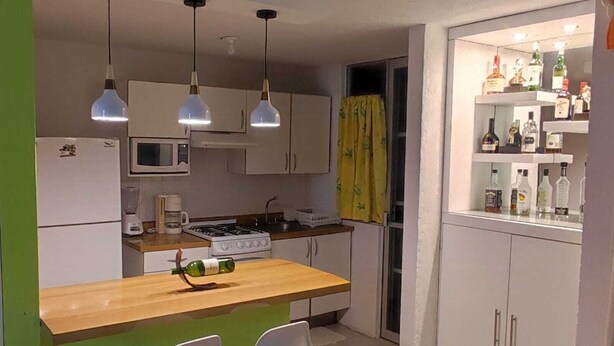 Eine kleine Küche mit einer Holz Arbeitsplatte, einem Herd, einer Mikrowelle und einem Kühlschrank. Zwei Pendelleuchten hängen von der Decke, ein gelber Vorhang ist angebracht und ein Regal enthält verschiedene Flaschen.