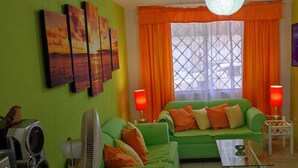 Ein Wohnzimmer mit einem grünen Sofa, orangefarbenen Kissen, einem Glastisch, einem Sony Lautsprecher und einem an der Wand befestigten Fernseher.