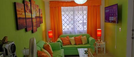 Una sala de estar con un sofá verde, cojines naranjas, una mesa de centro de vidrio, un altavoz Sony y un televisor montado en la pared.