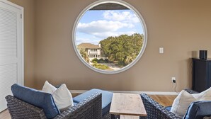 Habitación con ventanal circular que ofrece vista a una casa y árboles. Hay un sofá de mimbre con cojines azules y una pequeña mesa de madera.