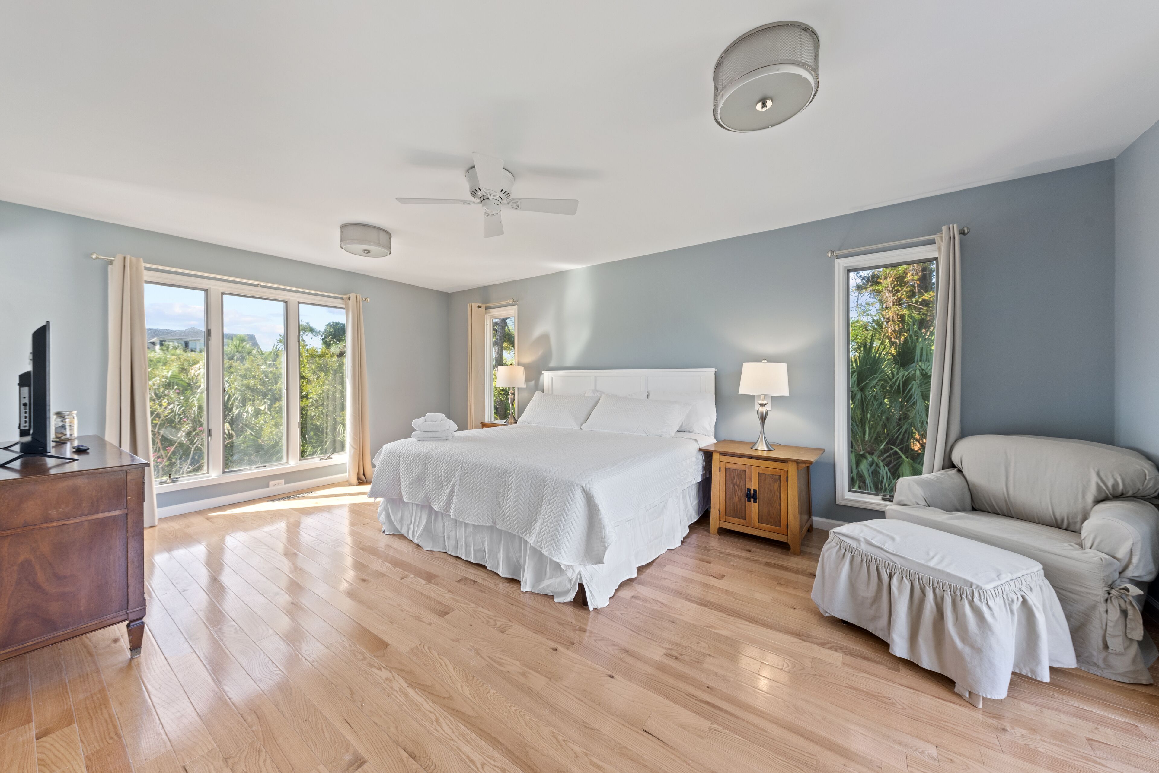 A spacious bedroom with a large bed, a wooden dresser, a nightstand with a lamp, a ceiling fan, and a view of the outdoors through large windows.