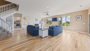Amplia sala de estar con pisos de madera, una escalera, una cocina al fondo y ventanas grandes.