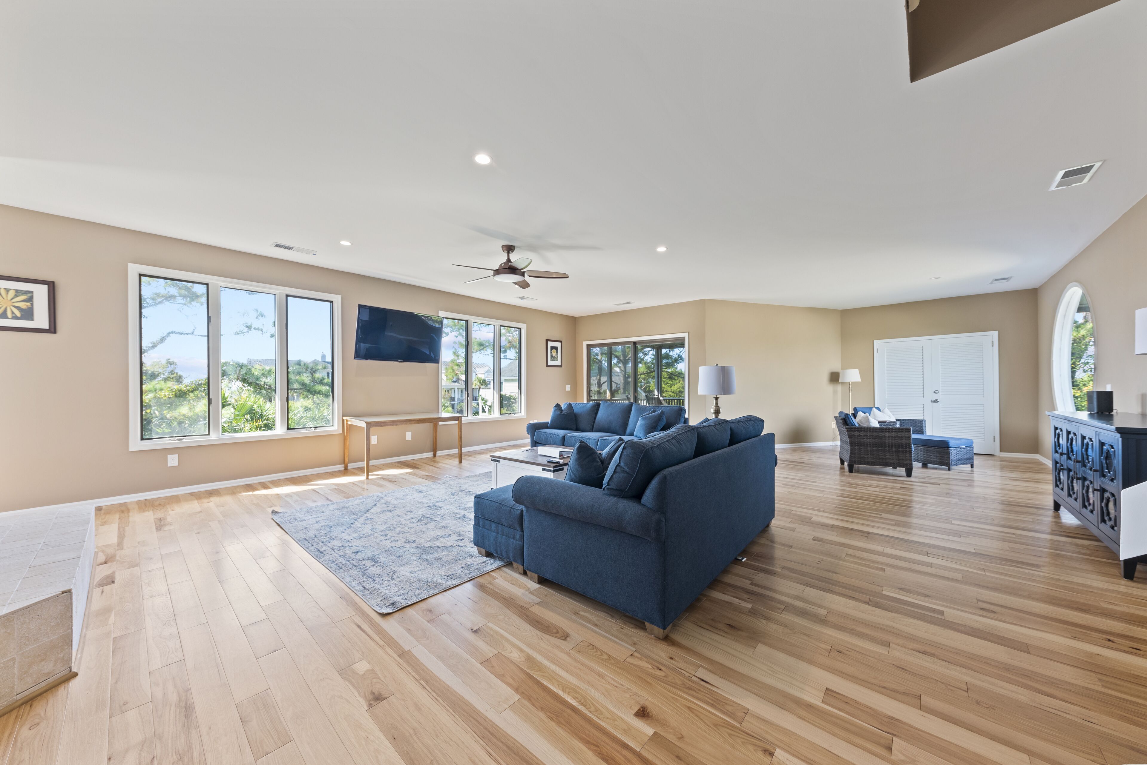 A spacious living room with a blue sofa, wooden flooring, large windows, and a ceiling fan.