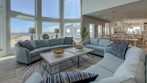 A spacious living room with large windows, a sectional sofa, a coffee table, and a view of the beach. - Royal Shell North | Oceanfront | Private Pool and Hot Tub (Virginia Beach)