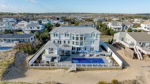 A large, multi-story house with a swimming pool, surrounded by a sandy beach and other houses. - Royal Shell North | Oceanfront | Private Pool and Hot Tub (Virginia Beach)