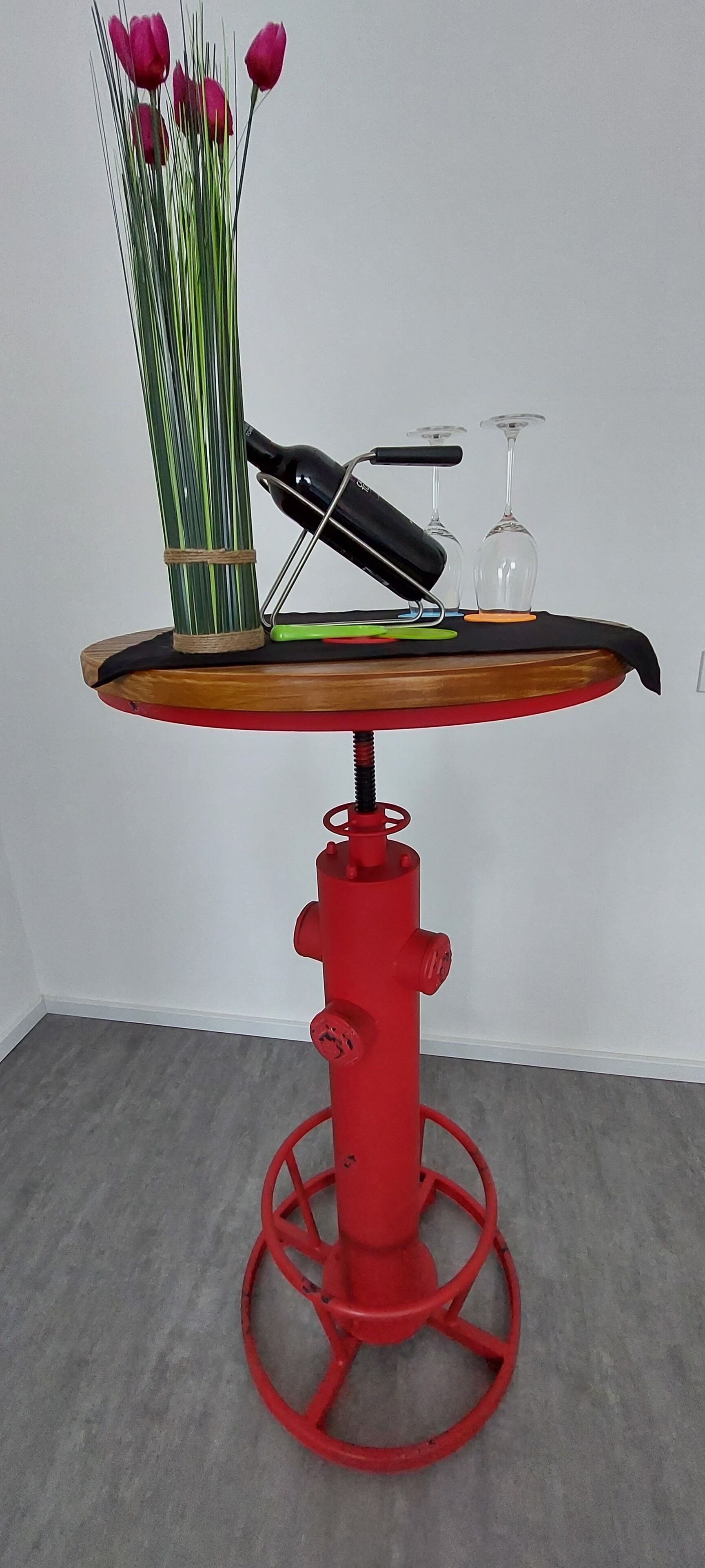 Een rode metalen tafel met een houten blad, een vaas met roze bloemen en twee wijnglazen.