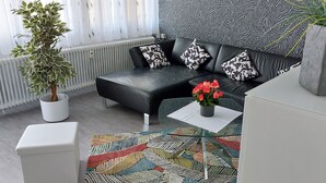 A modern living room with a black leather sofa, patterned cushions, a white side table with a vase of flowers, a colorful rug, and a potted plant. - Vacation apartment 90 m2, 2 minutes walk to the hospital in the center of Sulzbach  (Sulzbach/Saar)