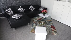 A modern living room with a black leather sofa, patterned pillows, a white side table, a glass coffee table, and a colorful rug. - Vacation apartment 90 m2, 2 minutes walk to the hospital in the center of Sulzbach  (Sulzbach/Saar)