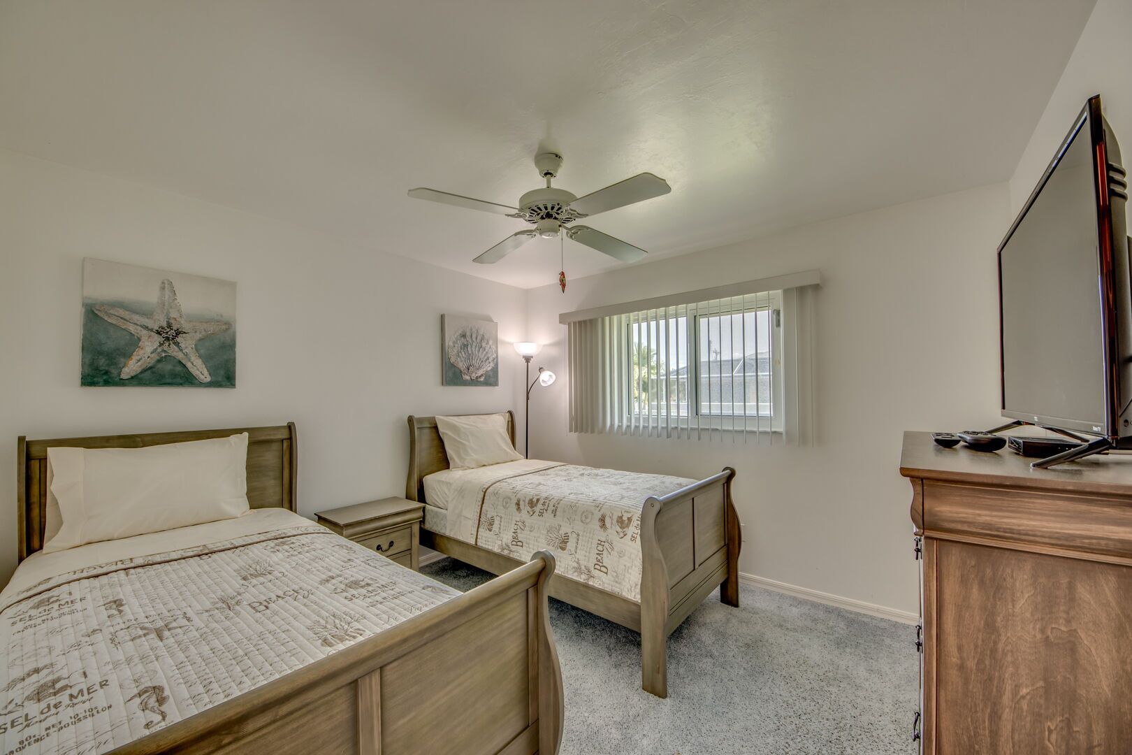 A bedroom with two beds, a ceiling fan, a window with blinds, and a TV on a wooden dresser.