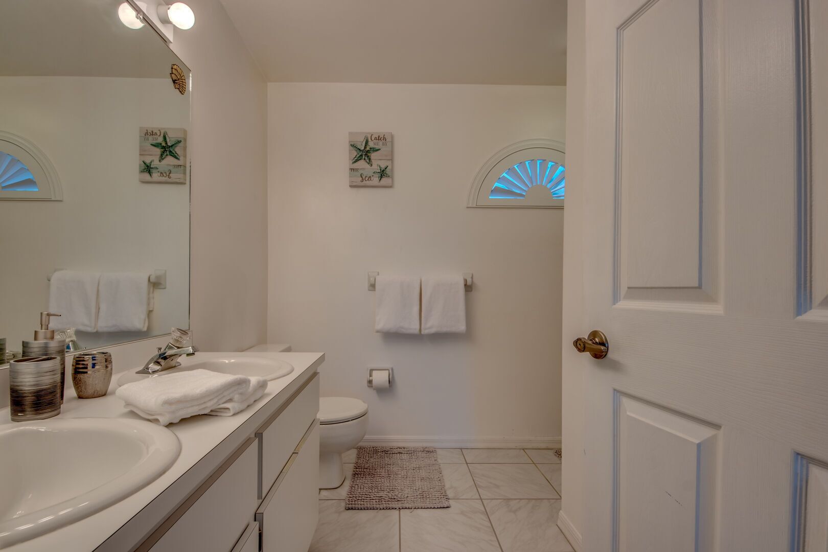 Un bagno con doppio lavabo, water e portasciugamani. Si notano decorazioni a parete a forma di stella marina e una ventola sopra il water.