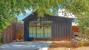 A rustic wooden shed with a large window, surrounded by trees and a paved driveway.