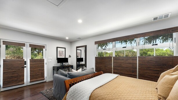 A bedroom with a large bed, a desk, and a chair. There are two windows with wooden blinds, and a door with a sliding glass panel.