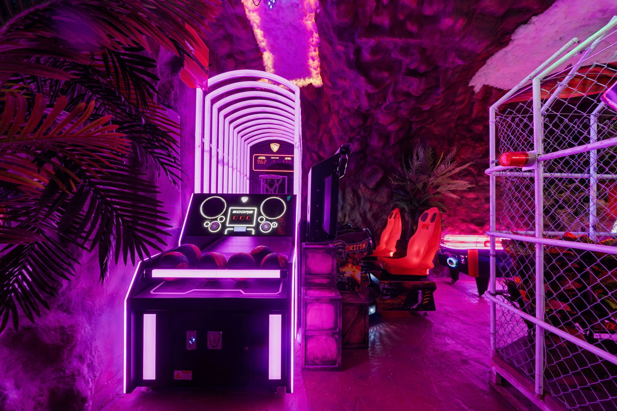 Un salón de juegos neón con una configuración de juego de carreras, una máquina de pinzas y una jaula con un objeto rojo.