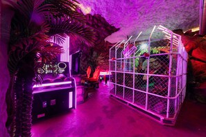 A room with a neon-lit arcade, a cage with a toy, and a tropical plant.