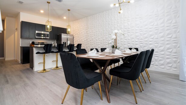 A modern kitchen with dark cabinetry, a central island, and a dining area with black chairs and a wooden table. - Luxury 3 Bed Home up to 8 guests- The Hub - 3236 TT (Kissimmee)