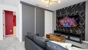 A modern living room with a black leather sofa, a wooden coffee table, and a TV mounted on a geometric-patterned wall. - Luxury 3 Bed Home up to 8 guests- The Hub - 3236 TT (Kissimmee)