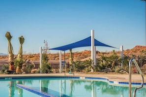 Ein Swimmingpool unter einem blauen Sonnensegel, umgeben von Wüstenpflanzen und blauem Himmel.