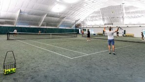 Indendørs tennisbane med net, en kurv med tennisbolde og et par personer der spiller.