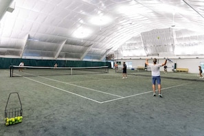 An indoor tennis court with a net, a basket of tennis balls, and a few people playing. - Sailors Rest | 844 ft to the beach (Corolla)