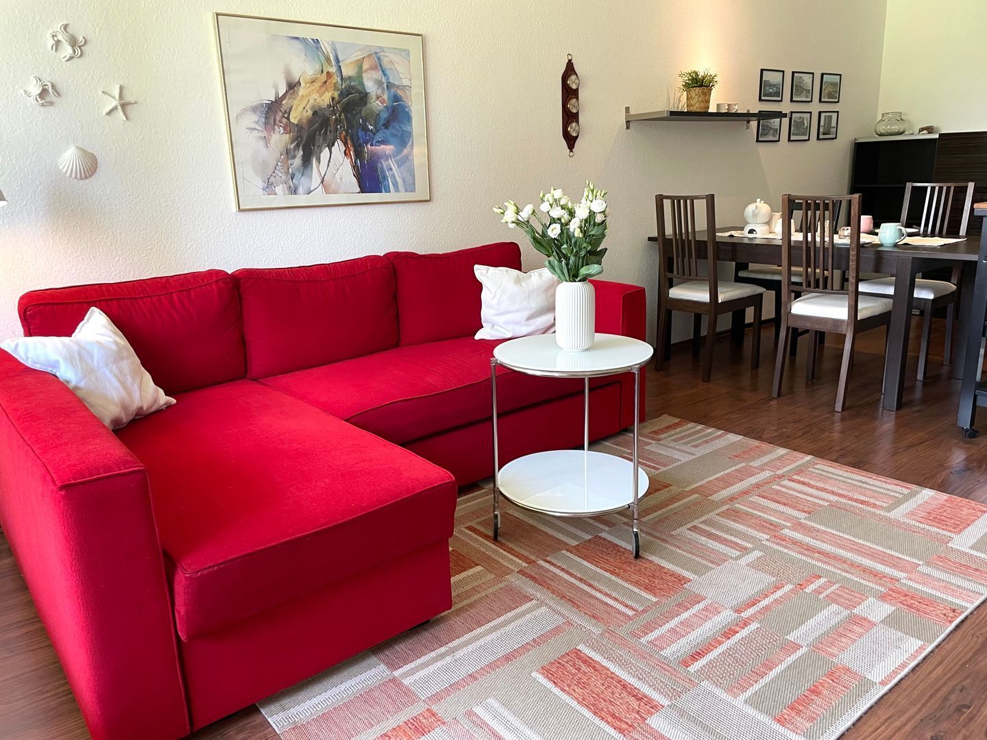 A red sectional sofa with a white pillow, a small white side table, a patterned rug, a dining area with chairs and a table, and a wall clock.