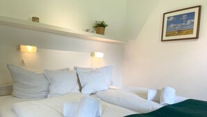 A neatly made bed with white linens and a green throw blanket, a wall-mounted shelf with a plant, and a framed picture on the wall.