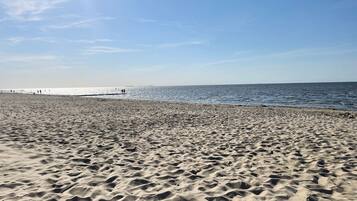 Een zandstrand met voetstappen die naar het water leiden, een heldere lucht en een pier in de verte.