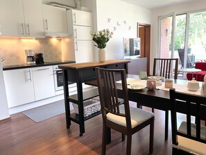 A modern kitchen with white cabinets, a wooden dining table, and a view of the outdoors through a glass door. - Fewo14 - Haus Land & Meer (Cuxhaven)
