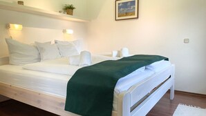 A single bed with white bedding and a green throw, a bedside table, a wall-mounted light, a framed picture, and a small plant on a shelf. - Fewo14 - Haus Land & Meer (Cuxhaven)