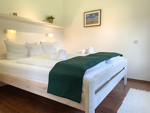 A single bed with white bedding and a green throw, a bedside table, a wall-mounted light, a framed picture, and a small plant on a shelf. - Fewo14 - Haus Land & Meer (Cuxhaven)