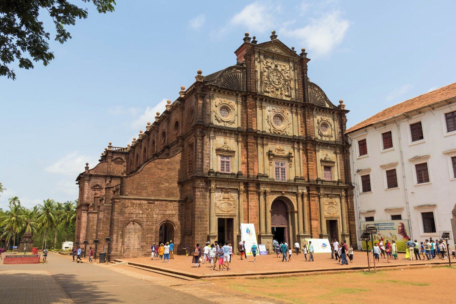 Uma igreja histórica com uma grande torre de sino e um relógio, rodeada por uma praça com palmeiras e pessoas.