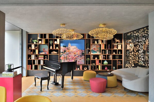 A modern living room with a grand piano, a colorful patterned rug, and a large bookshelf filled with books and decorative items. - Citadines Arpora Nagoa Goa (Arpora)