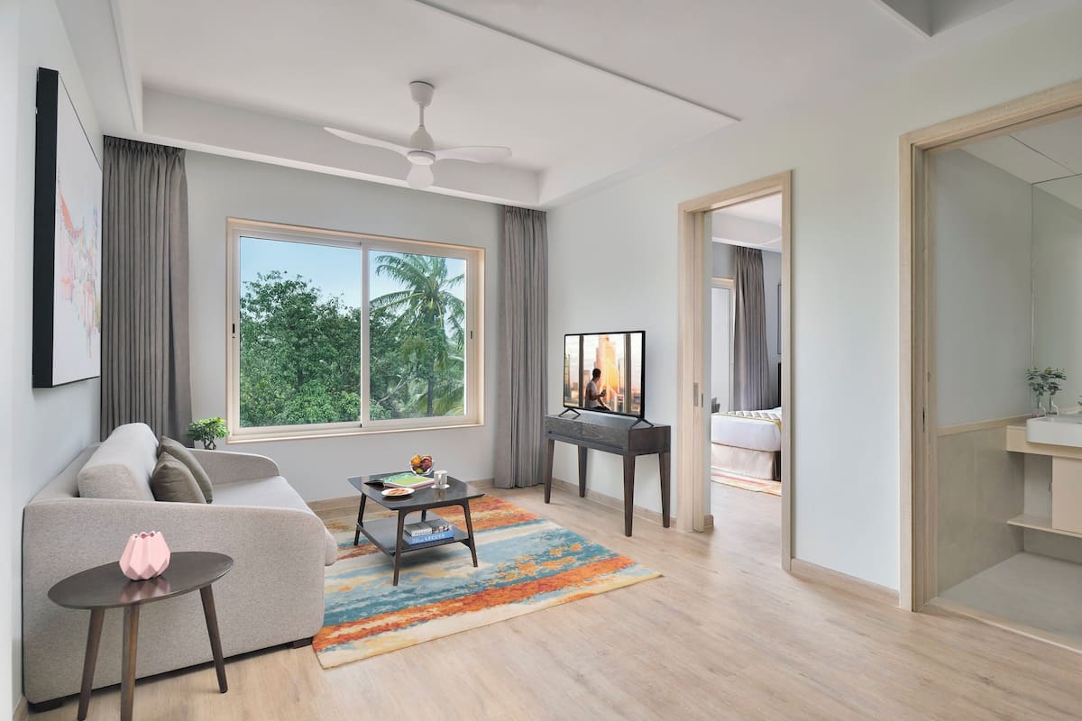 Una sala de estar moderna con sofá, mesa de centro y mueble para televisión. Hay un ventanal con cortinas, un ventilador de techo y vistas a árboles.