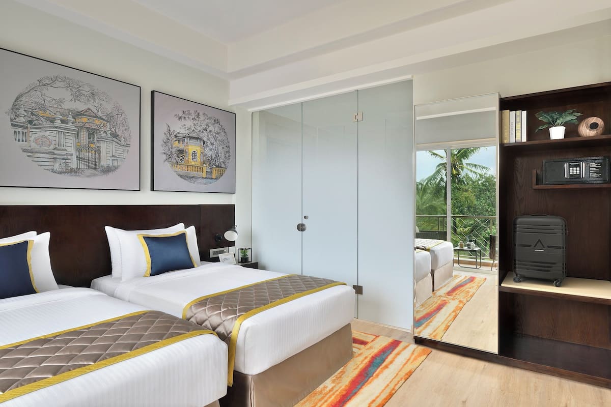 Habitación de hotel con dos camas, un armario, un escritorio con silla, una mesita de noche con lámpara y vistas al exterior.