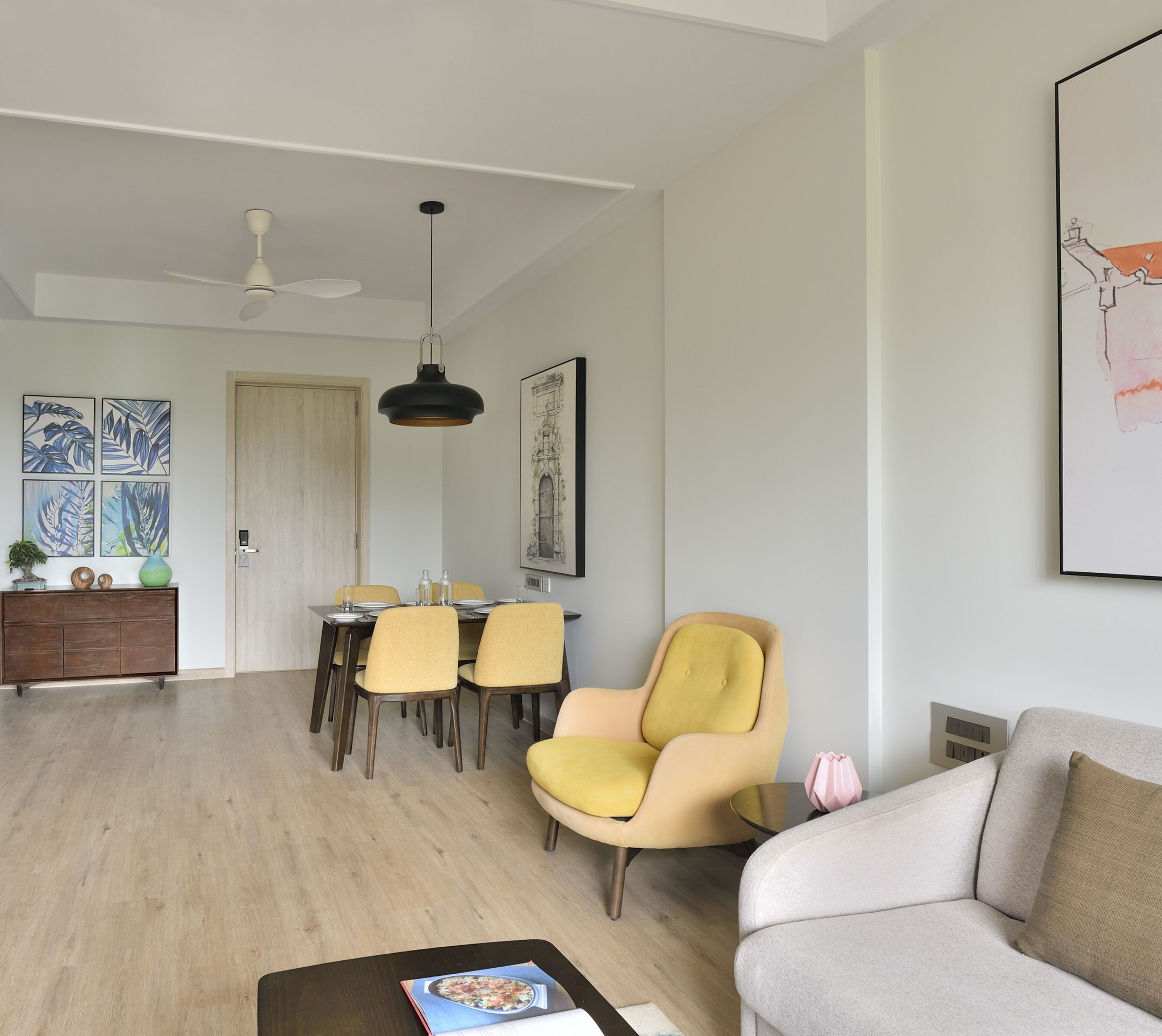 A modern living room with a sofa, a yellow armchair, a dining area with chairs, a ceiling fan, and artwork on the walls.