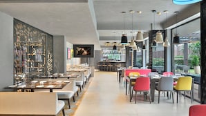 Un restaurant moderne avec de longues tables et des chaises colorées, un coin bar et de grandes fenêtres donnant sur l’extérieur.