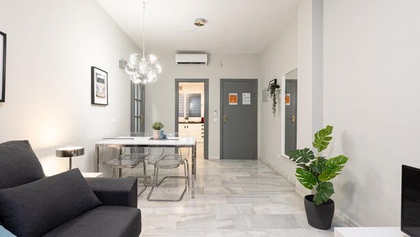 A modern living room with a grey sofa, a dining table with chairs, a potted plant, and a TV stand.