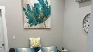 Una sala de estar con un sofá negro, dos cojines blancos y amarillos con estampado, un reposapiés beige y un cuadro abstracto color turquesa en la pared.