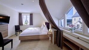 A modern hotel room with a large bed, a TV, a chair, a window with curtains, and a view of a building outside. - Ferienwohnung 10 für 1-5 Personen (Bernau am Chiemsee)