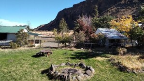 A rural landscape with a stone fire pit, a wooden bench, and a fenced area with a house and barn.