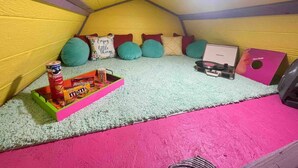 A small, cozy room with a sloped ceiling, colorful pillows, a small table with snacks, and a record player.