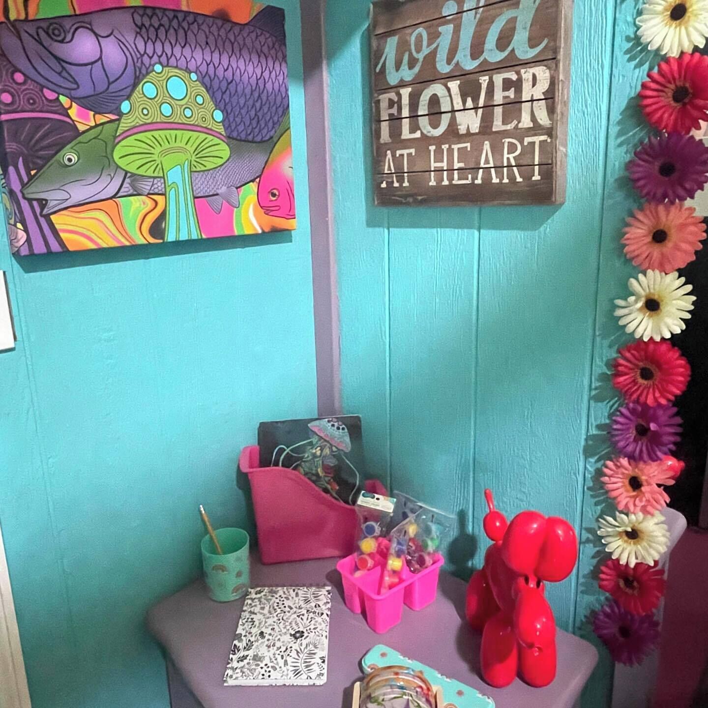 Ein raum in Türkis mit einem Schild mit der Aufschrift 'wild flower at heart', einem farbenfrohen Fischgemälde und einem Schreibtisch mit Notizbuch, Stift und dekorativen Elementen.