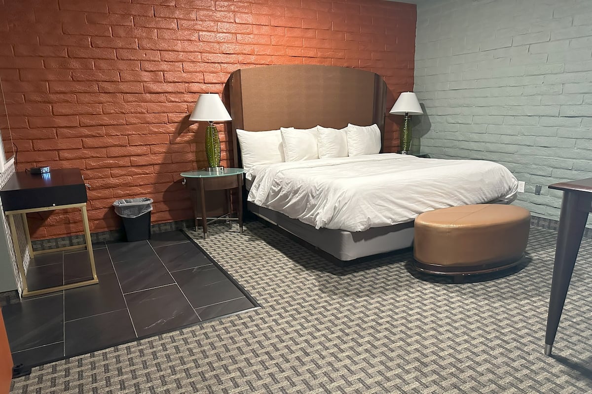 A hotel room with a brick wall, a bed with white bedding, a round ottoman, a small table, and two nightstands with lamps.