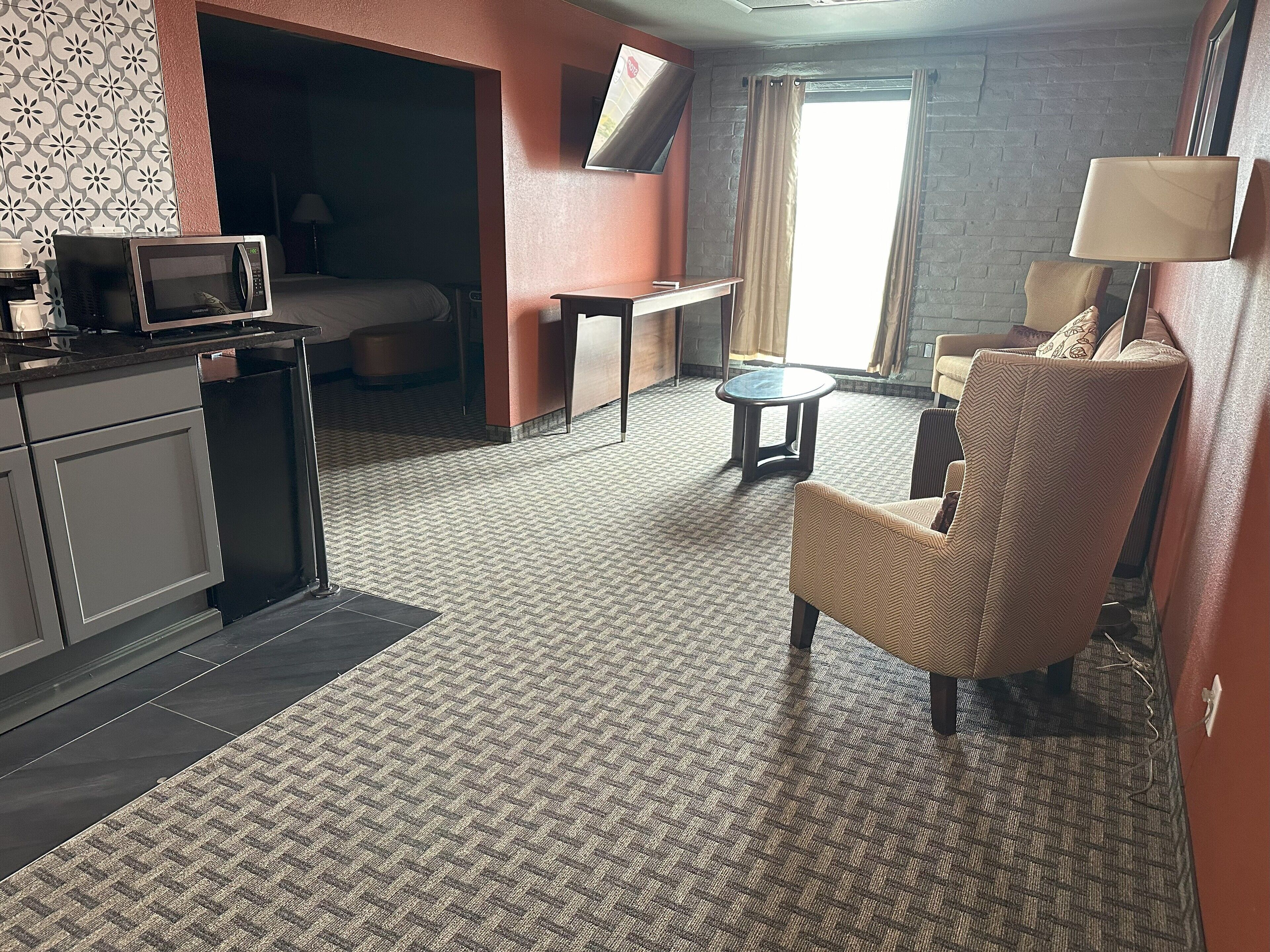 Habitació d'hotel amb petita cuina, un llit, televisió, escriptori, cadira i finestra amb cortines.