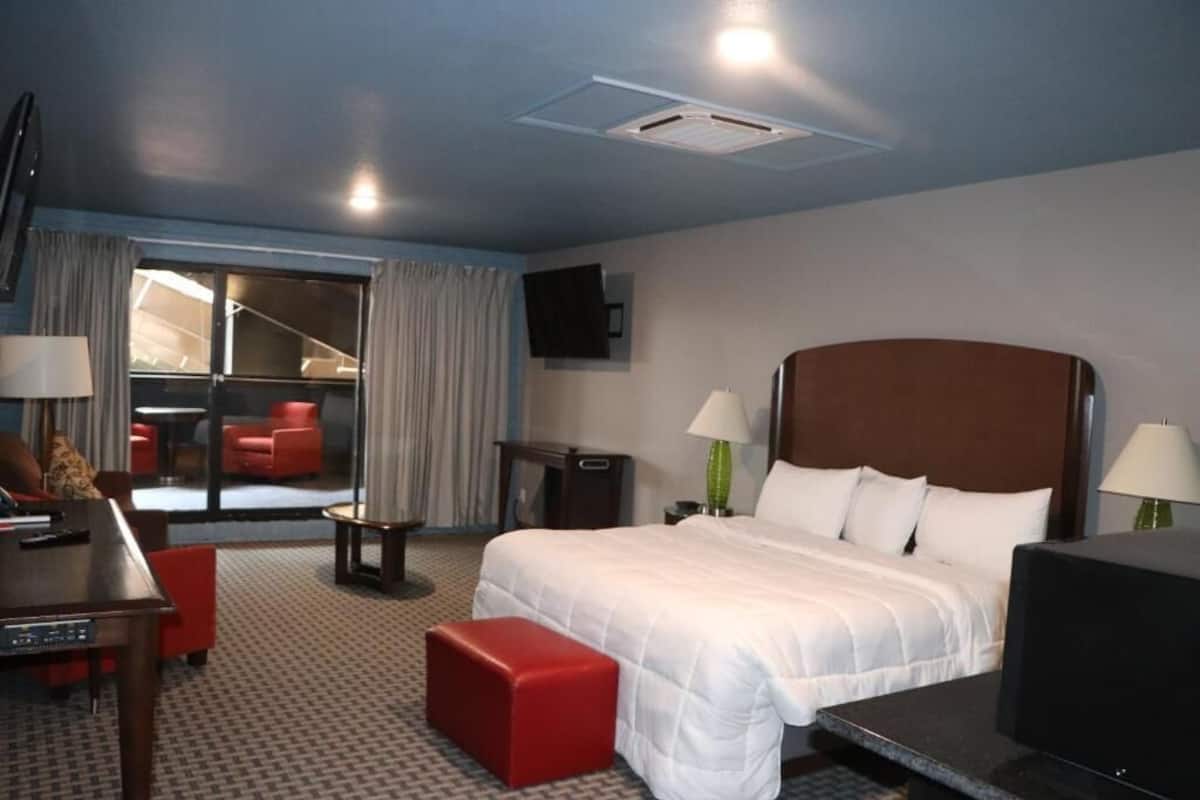 A hotel room with a large bed, a desk with a chair, a TV, and a seating area with a red chair and ottoman.