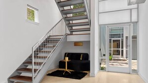 Un interior moderno con una escalera, un sofá negro, una mesita redonda y una puerta de cristal que da al exterior.