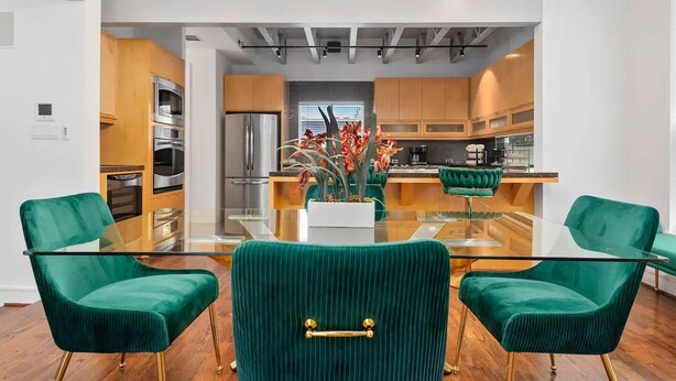 Una cocina moderna con electrodomésticos de acero inoxidable, armarios de madera y una isla central con cubierta de vidrio. Hay dos sillas de terciopelo verde con patas doradas alrededor de una mesa de vidrio.