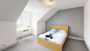 Une chambre avec un plafond en pente, un lit doté d’une tête de lit en bois, un bureau avec une chaise et une fenêtre avec des stores.